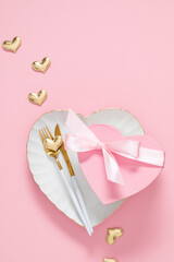 Romantic Valentine's Day setting: White heart-shaped plate with golden cutlery, pink heart gift box with ribbon, and gold hearts on pink background. Elegant flat lay for wedding or anniversary.