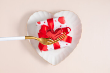 Romantic Valentine's Day concept: Small red heart on a golden fork, white heart-shaped plate, and a gift box with a red bow on a beige background. Elegant flat lay for wedding or anniversary.
