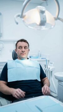 Vertical video of nervous man waiting in dentist chair. Anxious male patient with blue bib looking around fearfully before dental treatment. Dental phobia and fear concept