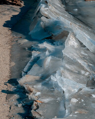 Vertical close-up shot of jagged ice slabs pushed onto a sandy shoreline during a winter freeze. The translucent blue ice blocks show detailed textures and cracks under bright daylight. © Denis Chubchenko