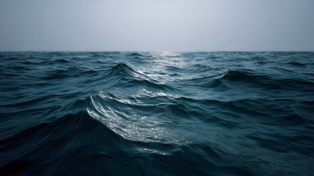 A moody deep blue ocean surface with moderate waves under a hazy overcast sky stretching to the horizon