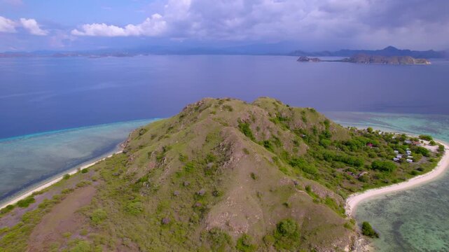 Backward-moving aerial footage revealing the peaks of Komodo National Park in Kanawa Island, Indonesia, a small tropical island surrounded by crystal-clear turquoise waters and coral reefs.