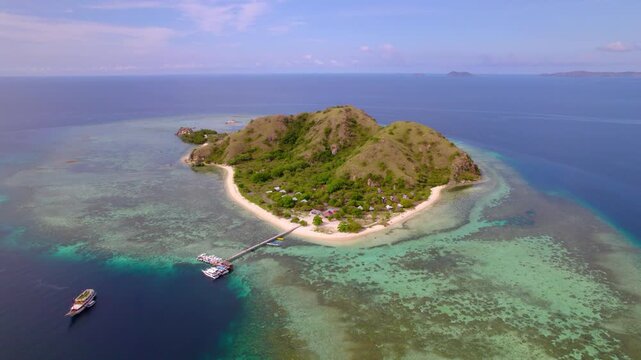Birds eye view of Kanawa Island in Indonesia showcasing private yachts and boats anchored near a wooden jetty, with small beachfront cottages and huts along a pristine white sand beach.