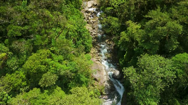Drone follows mountain river rapids through dense jungle and tilts up to show Cascada El Rocio Machay waterfall plummeting from high cliffs in lush tropical landscape of Banos Ecuador