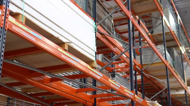 Close up of orange metal industrial shelves holding large stacks of wood panels and plywood in a professional lumber distribution center or hardware store warehouse.