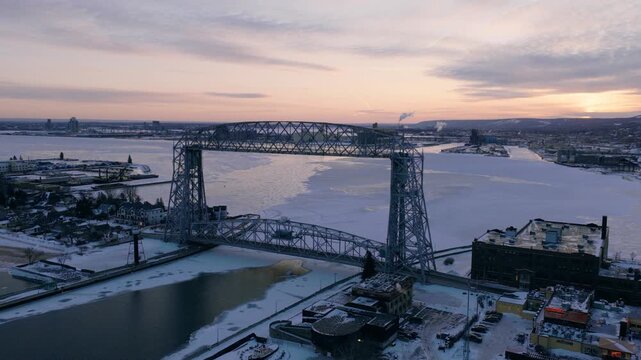 Wide drone shot of the Duluth Aerial Lift Bridge and frozen harbor during a vibrant winter sunset.