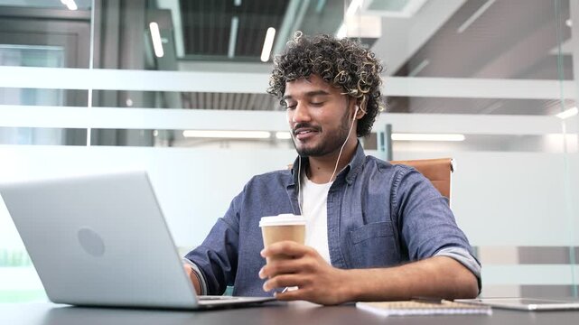 Happy young businessman working on a laptop holding a cup of coffee sitting at a workplace in business office. Smiling handsome male worker with headphones browsing web on computer or chatting online