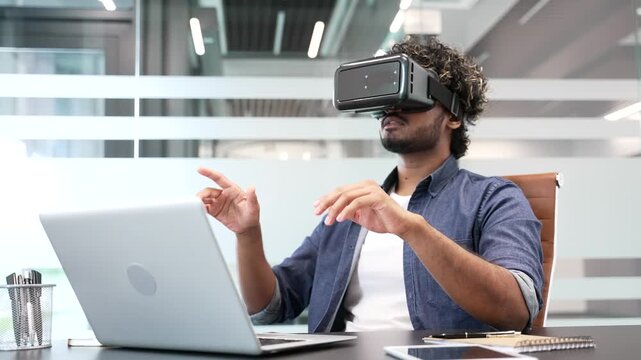 Young businessman works using VR glasses in virtual reality simulator sitting at workplace in modern business office. Worker uses gestures to control computer programs and flips through virtual pages