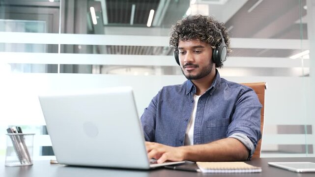 Focused businessman or IT specialist in wireless headphones works on a laptop sitting at a workplace in a business office. Serious coder developer is programming, busy with a project on the computer