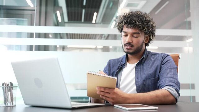 Young businessman watching video call conference or training notes in notebook looking at laptop screen sitting at workplace in business office. Entrepreneur or manager communicates remotely online