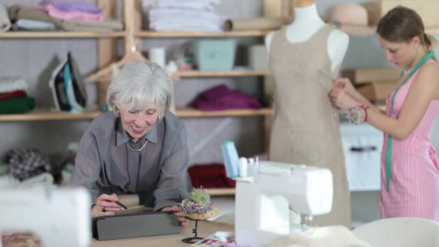 Mature dressmaker draws a design on an electronic tablet against the background of an assistant in a sewing workshop. High quality 4k footage