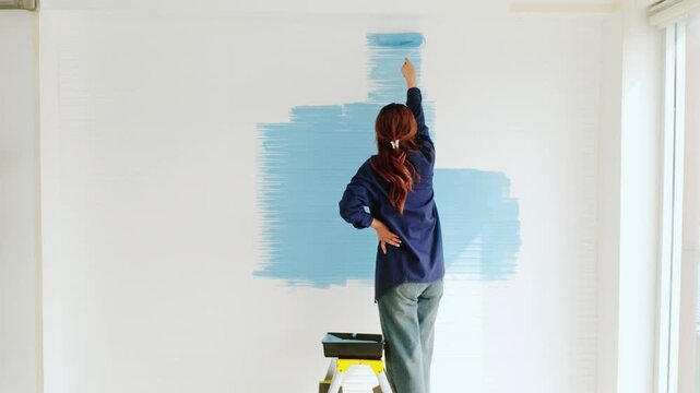 Person on a step stool painting a white interior wall sky blue during a DIY home renovation.