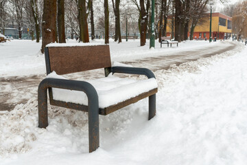 A wooden bench covered with fresh snow stands next to a plowed park path lined with bare winter trees. Urban public space, cold season atmosphere, and quiet outdoor leisure concept with copy space.