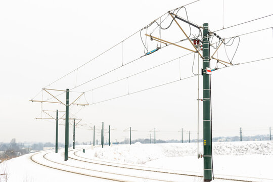 Curving railway track running through a snowy field with green catenary poles and overhead power lines. Minimal winter scenery highlights rail transport infrastructure, electricity supply, and cold-we