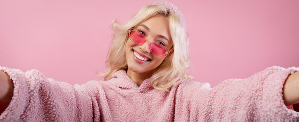 A woman stands against a pink background wearing pink sunglasses and a fluffy hoodie. She is smiling and appears to be taking a selfie, enjoying the moment. © Prostock-studio