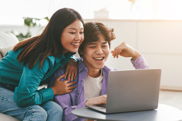 Fototapeta premium Millennial Asian couple in casual wear using laptop, having online meeting, watching interesting movie in living room. Young family communicating on web, surfing web together