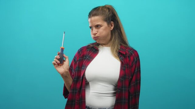 Woman holding a screwdriver upright, puffing her cheeks and squinting while wearing a red plaid shirt in studio; frustration doubt.