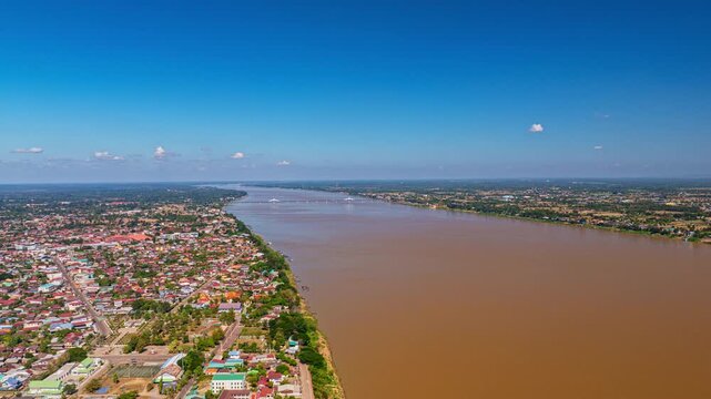 A dramatic aerial view of a riverside city illuminated by the warm glow of sunrise, with golden light spreading beneath a textured sky of thick clouds. The riverbank, buildings, and vibrant horizon 