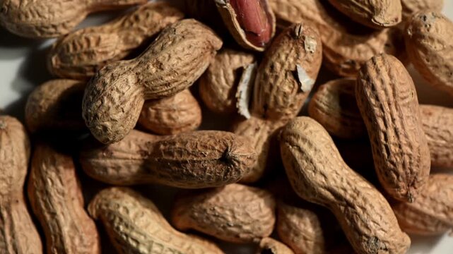 Aerial flat lay of roasted, golden-brown peanuts in their cracked-open shells spinning on a textured dark slate stone surface. Some shells slightly ajar revealing the nuts inside. Warm, earthy, rustic