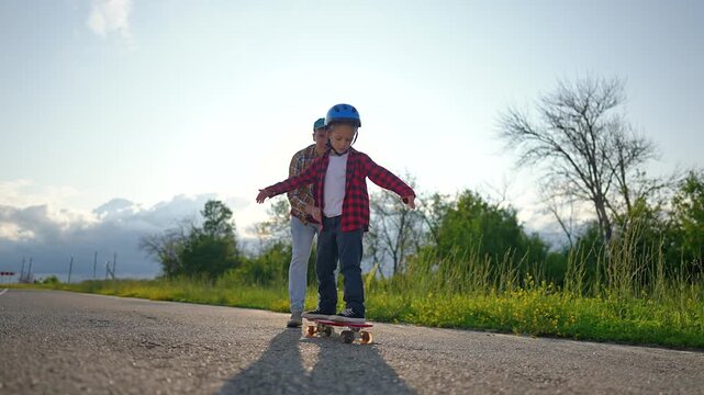 Father teaches son skateboarding on road outdoors. Dad helps child learn skating with helmet. Parent supports boy riding skateboard. Father teaches son on road. Boy learns skateboarding with dad.