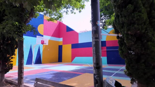 Dynamic POV walking shot approaching the brightly colored, modern street art basketball court in Jardines Huerto de Orive park, Cordoba, Spain.