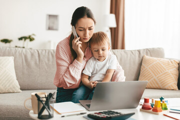 Millennial mother working on laptop computer and talking on phone, sitting on sofa with son on her...