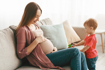 Happy pregnant mother sitting on sofa and touching her belly, spending time with her little child boy, playing together on sofa at home