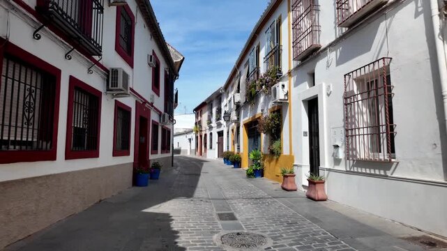 POV walking down a narrow, sunlit street lined with vibrant blue and red flower pots in the historic San Basilio neighborhood of C&oacute;rdoba, Spain.