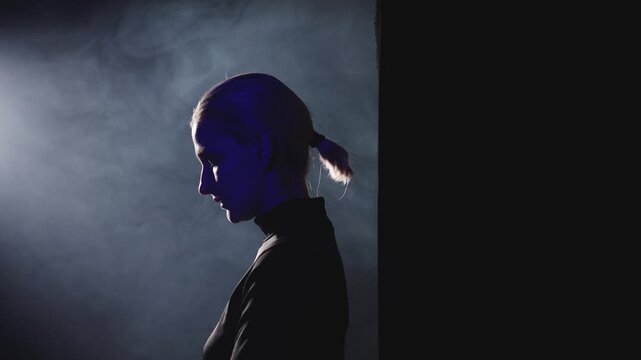 A young blonde woman with a short ponytail steps into a powerful side light. High-contrast video with her thoughtful profile illuminated, symbolizing the decision to stop being silent and embrace publ