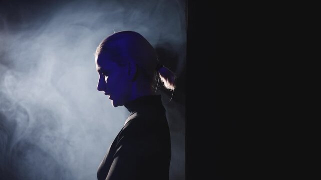 A young blonde woman with a short ponytail steps decisively into a bright side light. High-contrast video captures her profile, symbolizing the courage to speak out and overcome stage fright.