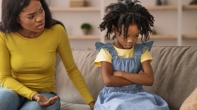A mother talks to her daughter who sits with arms crossed on a couch. The daughter looks upset and avoids making eye contact with her mother. The setting is a living room during daytime.