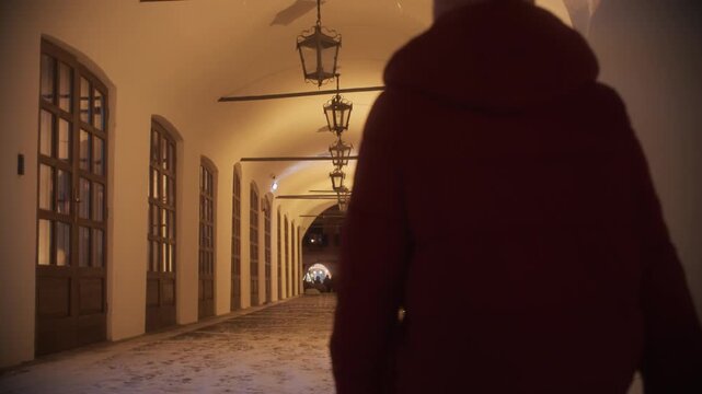 Woman in winter jacket and bobble hat walking through an illuminated archway corridor with snow on the ground at night