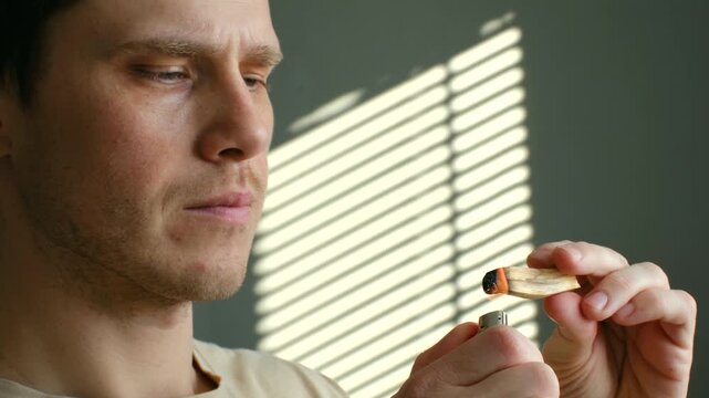 Pensive man lighting palo santo smudging stick with lighter for purification ritual, holding burning wood, watching aromatic smoke wafting through sunlit room. Man lighting palo santo stick for ritual