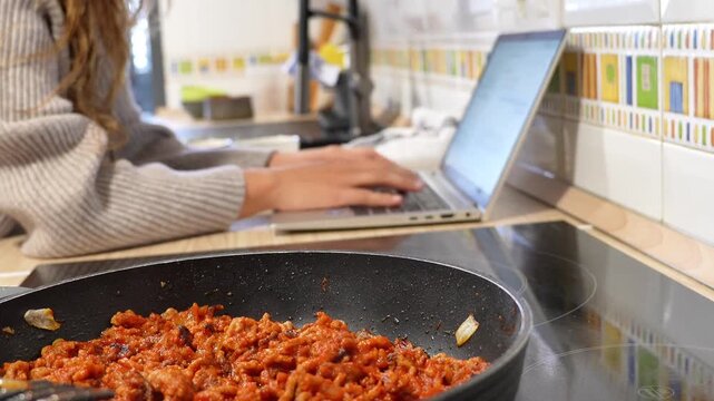 A woman is typing on a laptop while cooking food in a pan. The laptop is open and the woman is focused on her work. Concept of multitasking and productivity