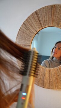 A woman is brushing her hair with a brush. The brush is white and the woman is looking at herself in a mirror