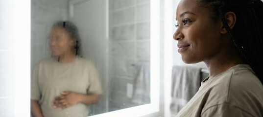Obraz premium Black woman looking in steamy bathroom mirror, reflection shows her hand on stomach, concept of pregnancy, selfreflection, and anticipation