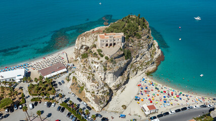 Aerial view of Sanctuary of Santa Maria dell'Isola in Tropea, Calabria, Italy. The ancient church stands on a rocky promontory, overlooking a crowded sandy beach filled with colorful sun umbrellas. © Stefano Tammaro