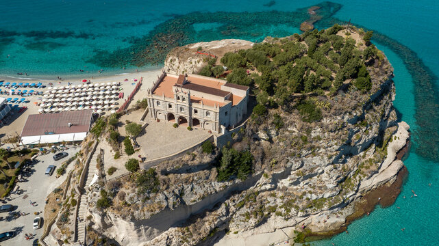 Aerial view of Sanctuary of Santa Maria dell'Isola in Tropea, Calabria, Italy. The ancient church stands on a rocky promontory, overlooking a crowded sandy beach filled with colorful sun umbrellas.