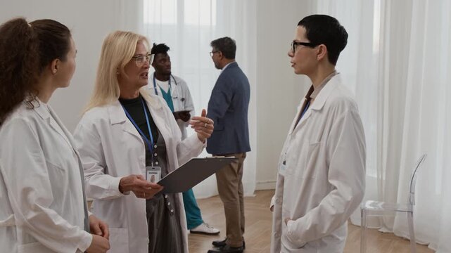 Brunette middle-aged doctor listening to blonde mature colleague holding clipboard, young nurse watching them standing nearby, two men chatting in background