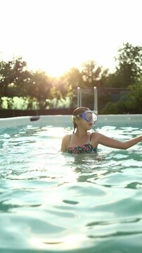 A teen girl dances and has fun in a swimming pool wearing a diving mask. The sun shines brightly, creating a joyful atmosphere. She enjoys the water and her time outdoors.