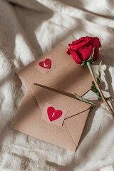 Two brown envelopes with red heart stickers sit next to a red rose on white fabric. Valentine's Day concept. 