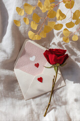 A red rose sits next to a letter with hearts on a light fabric surface. Dried leaves create a contrasting backdrop. Valentine Day concept.