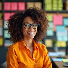 Naklejka premium Smiling woman with glasses in orange shirt. She has curly hair and works in a colorful office. This businesswoman is happy and confident at her desk, looking forward to success.
