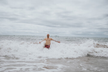 A shirtless man in red trunks stands in foamy surf, arms wide, smiling as waves crash around him. Overcast skies lend a cool, lively mood to a dynamic beach moment.