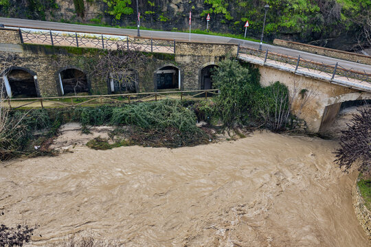 View of the surging, muddy river water rushing under the old stone bridge and adjacent arched building, Setenil de las Bodegas, Andalusia, Spain.