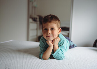 Fototapeta premium Smiling toddler boy resting chin on hands on white bed at home 