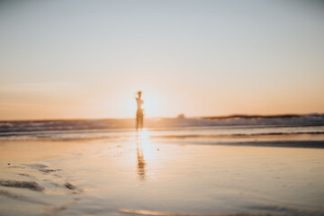 A lone silhouette walks along a serene shoreline as the sun sinks, casting warm golden light and long reflections on the wet sand. Ideal for travel, romance, and relaxation themes. © qunica.com