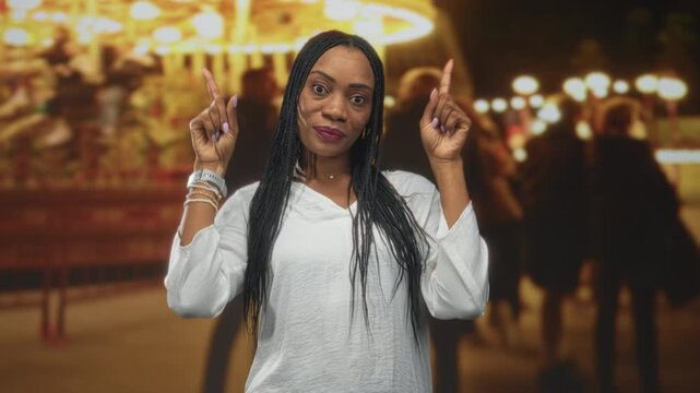 Woman in white blouse points index fingers upward beneath carousel and string lights on a street at night; celebration confidence.