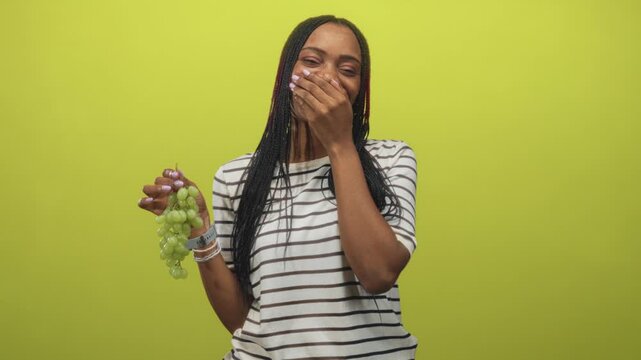 Woman holding green grapes, hand covering mouth laughing gesture, striped tshirt and bracelets visible in studio; joy playfulness freshness.