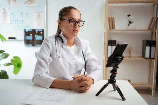 female doctor wearing eyeglasses and white coat using phone on tripod, consulting patient online. therapist making videocall in her medical clinic. Telehealth and telemedicine